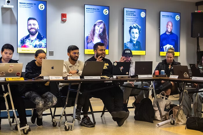 Student government members seated at a row of tables with laptops during a meeting, alumni portraits on the wall behind them